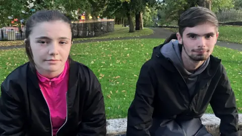 BBC Kenzie and John are pictured sitting on a wall. Kenzie has brown hair tied back and is wearing a pink zip up jacket and a black zip up jacket over the top. John has dark brown hair and is wearing a grey hoodie and a black waterproof jacket on top.