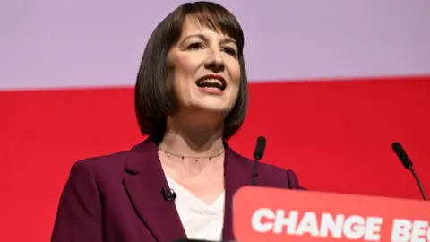 PA Media Rachel Reeves standing at a lectern with a maroon suit and white top