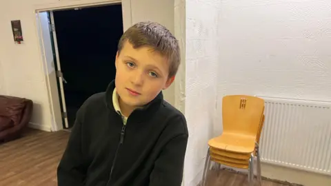 BBC/RICHARD EDWARDS Caiden is leaning on a table and looking straight into the camera. He has brown hair and is wearing a black top.