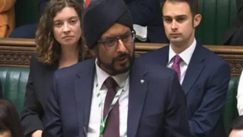 UK Parliament Warinder Juss, wearing a dark suit, stands in the House of Commons, flanked by other MPs sitting behind him