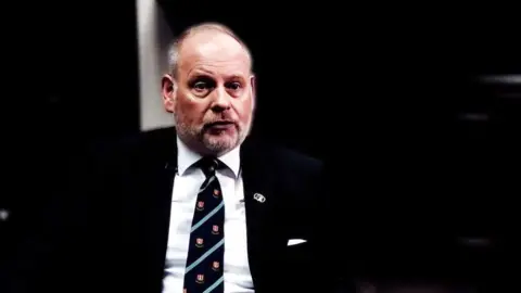 BBC Jonathan Nunn with short hair and a beard wearing a dark jacket and tie