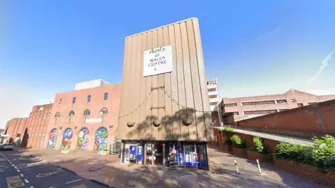 Google A large building with a ramp at the side of it. There is a sign which reads Prince of Wales Centre above a set of windows and glass doors. A section of the building set back from the entrance has a number of archways which each contain colourful murals.
