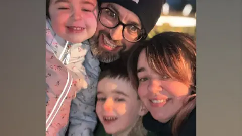 Cillian Kirwan Cillian Kirwan and his wife Louise and two children. They are all grinning at the camera. Cillian is wearing a black beanie, black glasses and his daughter is wearing a white floral coat, his son is grinning next to his mum who has long brown hair.