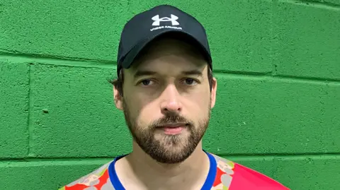 BBC A man in a baseball cap and pink t-shirt with a beard standing in front of a green brick wall