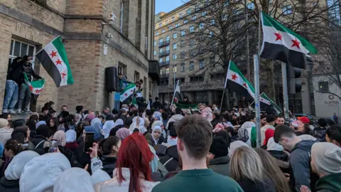 Large crowd of people waving Syrian flags outside Broadcasting House in Belfast, some men have climbed the building and have a large speaker... Clayton hotel and BBC building visible