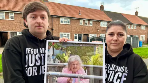 BBC Tristan Essex and Claire Stockdale are standing outside a row of terraced houses and holding a large framed photo of their grandmother. They are wearing black hoodies printed with white text, which reads: "Regulation and justice for Jessie".