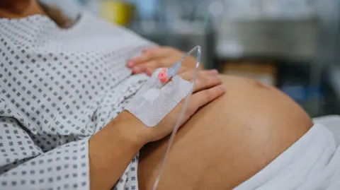 Getty Images Close up of pregnant woman’s belly. Woman in labour waiting for examination in maternity ward. Woman during early stage of childbirth at the hospital holding her belly, feeling painful contractions. She's wearing a hospital gown.