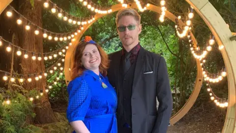 Handout Kelly Stuart and Nick Wilson. Kelly has red hair and wears a blue dress, while Nick is blonde and wears a suit. They are surrounded by trees and a string of lightbulbs is draped around a wooden structure behind them.