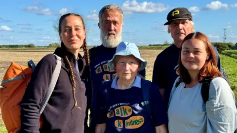 Mike Liggins Three women, including Cecile Roberts, and two men, wearing rucksacks, and some with blue "Walk of Kindness" T-shirts. pictured in a field on their charity walk. There is a blue sky and white clouds behind them.
