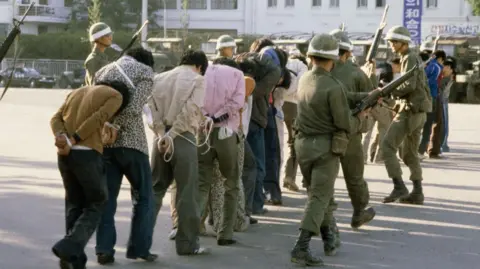 Getty Images Armed South Korean troops wearing helmets and with large guns round up protestors - mainly men, with their hands tied behind their backs, all in civilian clothes - following a raid in the city of Gwangju after an uprising against the Chun Doo-hwan dictatorship, South Korea, 27 May 1980