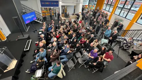 Claire Hamilton/BBC An aerial view of a large group sitting on fold up chairs facing a small stage inside the library