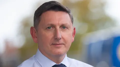 Guernsey Post A man with dark hair and wearing a light short and tie looks into the camera.
