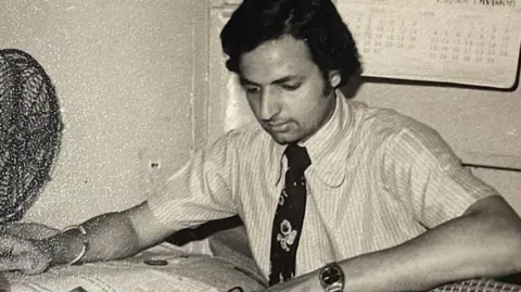 Handout Rajinder Verma in a black and white picture of him as a young man, sitting at a desk with a newspaper. He is wearing a short-sleeved shirt with a tie, a bracelet and watch, and has fan to one side of the table. There is a calendar behind him on the wall.
