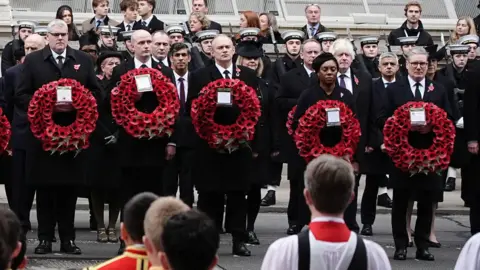 PA Media UK party leaders Gavin Robinson, Stephen Flynn, Ed Davy, Kemi Badenoch, and Sir Kier Stammer standing in a line holding poppy wreathes.