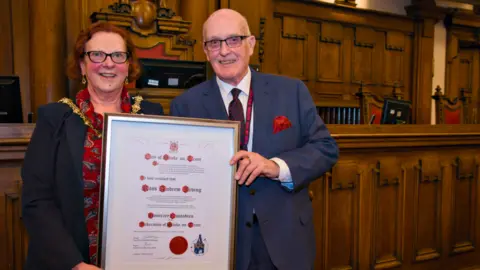 Stoke-on-Trent City Council Lord mayor of Stoke-on-Trent Lyn Sharpe poses with Ross Irving. Both are smiling, stood in the council chamber holding a framed certificate which recognises Mr Irving's appointment as the city's honorary custodian.