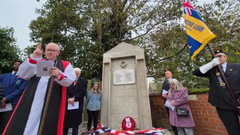 Stuart Woodward/BBC A war memorial in Shrub End near Colchester is rebuilt and unveiled. The Bishop of Chelmsford reads a short sermon. Standing next to the memorial is Sir Bob Russell, High Steward of Colchester, and Sue Starling, widow of Kevin Starling who campaigned to have the memorial re-built before his death in January 2024