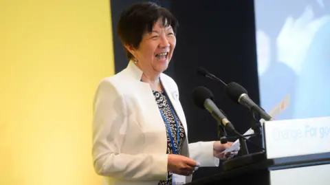 Pacemaker Anna Lo laughing during an Alliance manifesto launch.  She is wearing a white jacket, a monochrome patterned top and is speaking at a lectern.