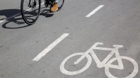 Getty Images A cycle lane with a bike painted on the floor in white paint with the wheels and pedals of a bike seen on the other side of the lane.