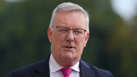 PA Media Mike Nesbitt, who has short grey hair, is standing outside. There are green trees behind him. He is wearing a navy suit, white shirt and pink tie. He is wearing glasses.