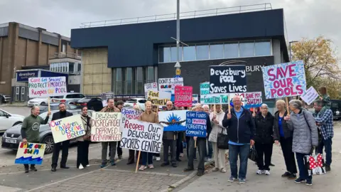 Olivia Richwald/BBC A group of men and women, outside a large building, holding placards with messages campaigning for the reopening of the leisure centre. The closed building can be seen in the background.  