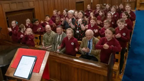 North Yorkshire Council Children sit singing in church pews, encouraged by two men and two women sat among them.