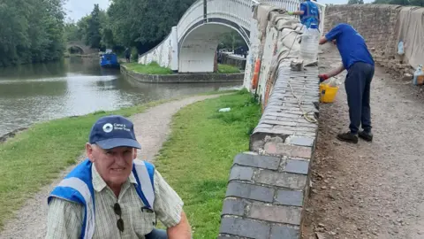Canal & River Trust A male volunteer in a blue cap reading 'Canal & River Trust' smiling at the camera. He is crouching down next to the bridge, where other people are working on repairing the bricks.