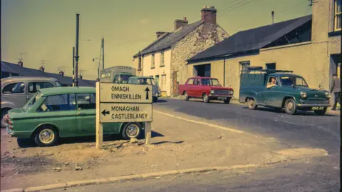 Armagh County Museum A digitalised photo from a film of street signs. Green, red and blue cars can be seen parked along the side of a small street. Looking west from Irish Street corner several cars are at the junction near the camera and the roofs of the terrace of new houses a partially visible in the background. An old and partially derelict house is on the right of the frame and a road sign reads "Omagh Enniskillen, Monaghan Castleblaney".
