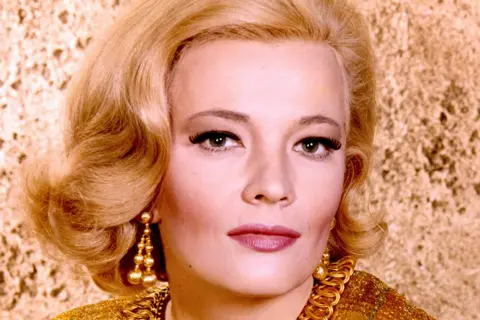 Getty Images Gena Rowlands photographed against a wall in what appears to be a publicity shot, taken around 1960 when she was 30. She is wearing make-up and staring straight at the camera, she has carefully styled shoulder-length blonde hair and has gold earrings which (from what little is visible) appear to match the colour of her dress.