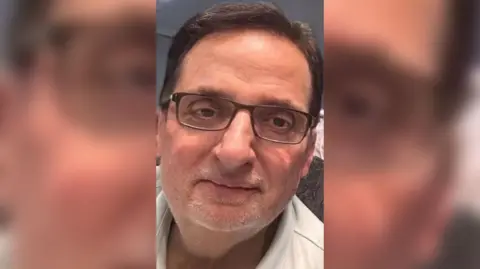Handout Rajinder Verma wearing glasses and a white shirt, with dark hair and a grey beard, leaning slightly to one side and looking at the camera.