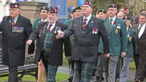 Pacemaker People in green and tartan uniforms wearing medals marching.