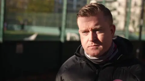BBC Eamon Gately in a black coat. He stands in front of a football pitch.