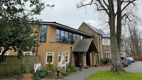 St Cuthbert's Hospice A brick building with an archway above the entrance. The windows on the ground floor have white frames, while those above have black frames and tinted glass. A large tree is growing at the front of the property close to where three cars are parked.