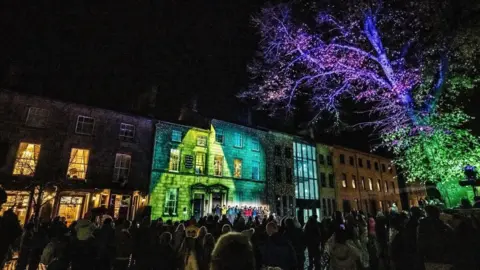 Robin Zahler A crowd of people in a Lancaster at night in front of a building lit up with green and blue lights and a tree lit up green and purple