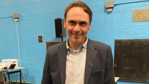 LDR A man standing in front of a blue wall in a hall. He has short brown hair, a striped shirt and a suit jacket.