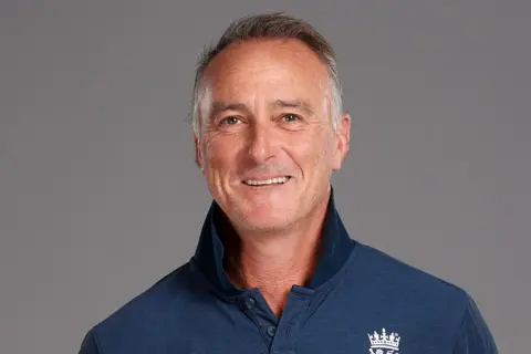 Getty Images Graham Thorpe is pictured against a gray background - he has grey receding hair and a lively smile. He is wearing a dark blue long-sleeved polo top with what looks like the England logo .