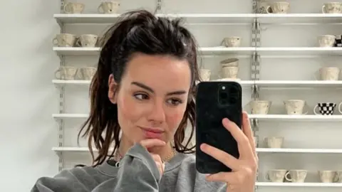 Mollie Warrington A young woman with dark hair, pulled back into a high ponytail, wearing a grey sweatshirt takes a mirror selfie, behind her is shelving filled with handmade neutral mugs.