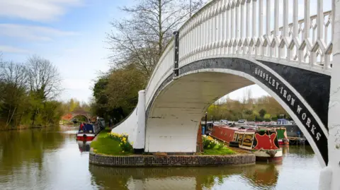 Canal & River Trust A white bridge with two red and orange boats sailing alongside it in the canal.