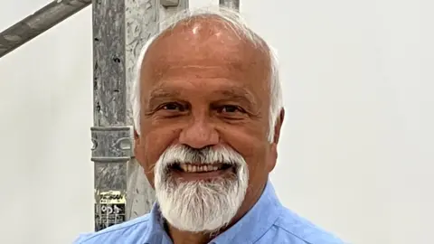 A man with a white goatee beard and a blue shirt standing in front of some scaffolding in a white painted room