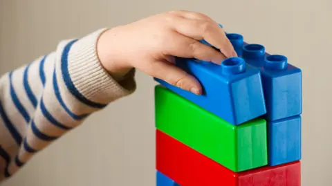 PA Media A child's hand reaches for toy building blocks. The child, wearing a grey sweatshirt with blue horizontal stripes, is placing a blue block on top of green red and blue blocks. The wall behind is a neutral colour.
