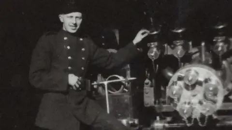 William Cruickshank A black and white picture of William Crocket standing on a piece of machinery. He is wearing a dark jacket with buttons on either side.