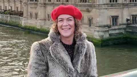 Family handout Sharon Doherty is wearing a red beret and fur coat.
She has short curly hair and is standing beside a river with a historic-looking building in the background.