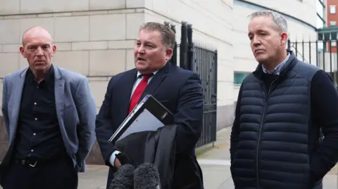 Pacemaker Photo outside court building showing three men in front of a microphone. Ciaran Shiels, centre, is wearing a navy suit and red tie.