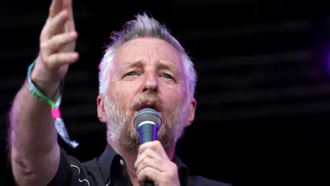 Getty Images Billy Bragg on stage holding a microphone in one hand and his other hand raised. He has white hair and a brown beard.
