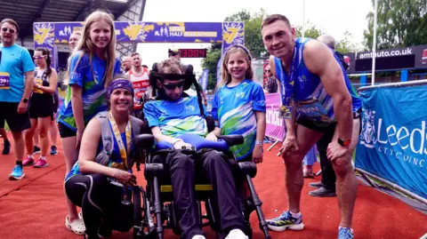 PA Media Group picture of Rob Burrow, his wife Lindsey, daughters Macy and Maya and Kevin Sinfield on a red carpet finish line after the Rob Burrow Leeds Marathon.