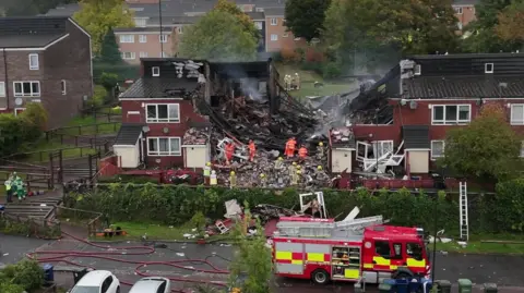 BBC Firefighters on site at the explosion. The houses in the centre have been ripped apart with debris on the ground. It is now daytime.