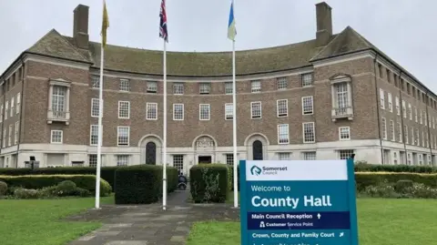 Daniel Mumby Somerset Council's County Hall pictured on a cloudy day. The town hall is in the background of the image. In the foreground is a sign which says 'Welcome to County Hall'. There are three flags poles outside of the building.