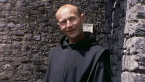 Father Thaddeus Kotik stands in front of the walls of Pembroke Castle. He is wearing a black habit. He is bald, with a little dark hair on the sides of his head. He is smiling into camera.