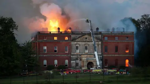 Getty Images Fire engines surround the house which is seen from the distance with large flames coming out of its left hand roof and also seen through lower windows on the right side of the building.