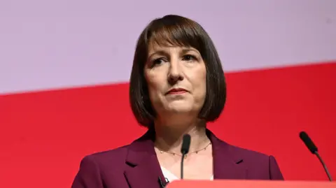 BBC Rachel Reeves with a thoughtful look on her face standing behind a podium during a speech. She is dressed in a purple jacket.