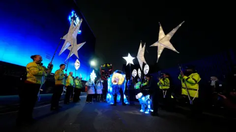PA Media Staff wear hi-vis jackets holding lit up star lanterns, with the bear lantern in the middle of the crowd 